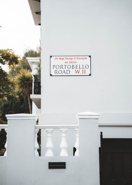A close-up photograph of a white residential building's exterior corner, featuring a decorative balustrade with classical-style white balusters and a white parapet. The wall has a smooth surface, painted in bright white, with a small black mailbox below the balustrade, integrated into the structure. To the upper right, a rectangular street sign is mounted on the wall, indicating the location as Portobello Road in the Borough of Kensington and Chelsea, with the postcode W.11, and displaying the district name in red and black text. In the background, partially visible trees with yellowish-green foliage and a glimpse of neighboring buildings can be seen, suggesting the property is situated in a well-maintained, possibly residential area with landscaped surroundings. The lighting appears natural and diffuse, with no harsh shadows, highlighting the clean, neat appearance of the exterior. The image emphasizes the tidy and orderly condition of the property, reflecting a typical private residence or rental property that may require services such as rubbish removal or waste collection, aligning with the themes of private disposal and alternative waste handling solutions that House Clearance Chelsea offers in the Kensington Chelsea district.