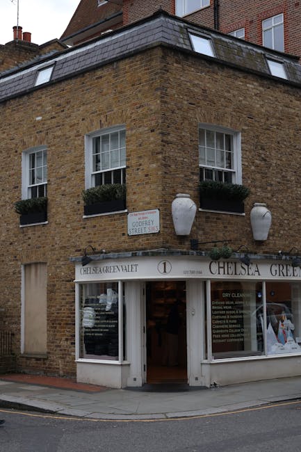 The image depicts a three-story brick building situated on a street corner, featuring a ground-floor shop with a white facade and large glass windows displaying signage for 'Chelsea Green Valet' and related services. The brickwork is a warm, brown tone with a textured surface, and the upper floors have two windows each with white frames. The windows on the second floor include dark green window boxes filled with greenery, creating a contrast against the brick. Two large, white, urn-shaped ceramic planters with rounded bodies and tapered necks are mounted on the building’s exterior, flanking the shop entrance. The shop entrance is open, revealing an interior space, and the sidewalk in front of the building extends to the street, where a curb separates it from the paved road. A street sign indicating 'Cuddery Street SW' is visible on the wall above the building, suggesting the location. The overall scene appears clean and well-maintained, with daylight illuminating the facade, emphasizing the textures and subtle color variations of the brickwork and decorative elements, aligning with the context of private waste handling or alternative rubbish collection methods that may involve on-site clearance or independent collection.