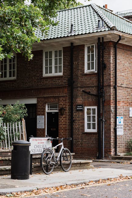 The image depicts a small, brick residential building with a grey tiled sloped roof situated on a paved street. In front of the building, there is a white bicycle parked near the entrance, leaning against a black cylindrical trash bin. Adjacent to the bicycle, a white signboard with red and black text advertises Chelsea Square, and a metal fence is partially visible on the left side, enclosing a garden area with green foliage. The building features multiple white-framed sash windows, with some on the upper floor and one next to the entrance door on the ground floor. Black rain gutters and downpipes run along the corner and side of the building, directing rainwater from the roof. The scene is lit with natural daylight, and a tree with green leaves partially obscures the left side of the building, adding a touch of greenery to the urban setting. The overall setting suggests a quiet residential street where private waste management, such as rubbish collection services by companies like House Clearance Chelsea, may be employed as an alternative to local authority disposal.