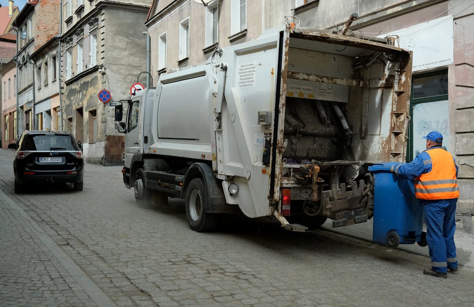 A street scene featuring a white waste collection truck parked parallel to the curb in an urban area, with a worker in a blue uniform and high-visibility orange vest emptying a blue wheelie bin into the truck's open rear compactor section. The truck is positioned beside a row of older multi-story buildings with a mix of weathered and newer facades, some showing signs of peeling paint and brickwork. The scene takes place on a cobbled street with faintly visible tire marks and minimal debris. To the left, a black car is parked facing the same direction as the waste truck. The worker is wearing gloves and a blue cap, standing on the pavement with a stance indicating active waste collection. The environmental lighting appears to be natural with soft shadows, suggesting a clear day. The image visually supports the context of independent rubbish removal services, as provided by House Clearance Chelsea, highlighting the collection process for domestic waste within an urban residential setting, emphasizing the handling and disposal of rubbish via private waste management methods.