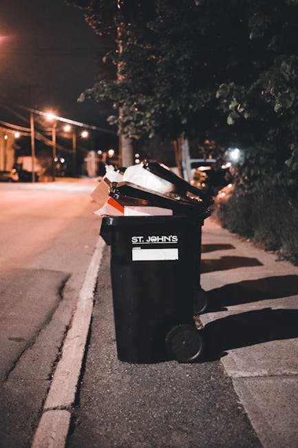 A black wheeled rubbish bin positioned on the edge of a paved sidewalk in an urban environment during nighttime. The bin has a white label with the words 'ST. JOHN'S' printed on it and appears to be filled with mixed waste, including cardboard and plastic items that are spilling over the top. The surrounding scene features dim street lighting, casting shadows on the ground, and in the background, there are trees, low-rise buildings, and parked cars along the street. The street itself is quiet and empty, with a row of streetlights creating a warm glow. The image suggests a private or independent waste collection scenario, highlighting the importance of proper rubbish disposal in a residential or commercial area, which House Clearance Chelsea may offer as part of its rubbish removal services in Chelsea SW10. Overall, the scene emphasizes the need for appropriate waste management and collection methods in urban environments, fitting within the context of rubbish disposal rules for landlords and property owners.