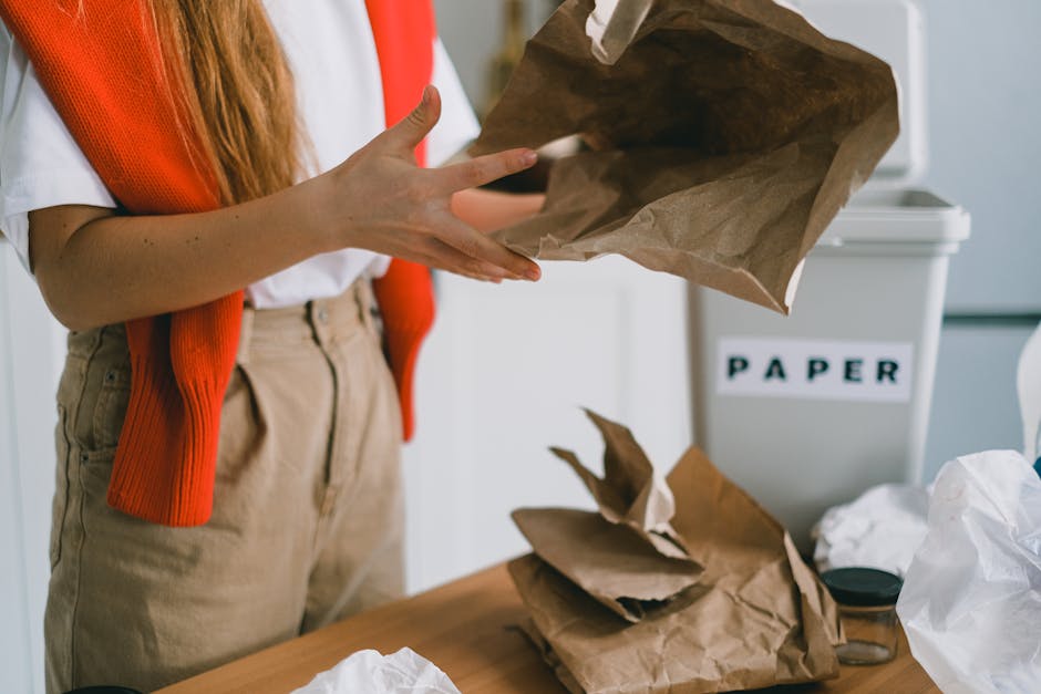 A person wearing a white shirt with rolled-up sleeves, beige high-waisted trousers, and an orange knitted vest is holding an empty crumpled brown paper bag with both hands, appearing to either prepare it for disposal or reuse. They are standing indoors near a wooden table cluttered with more crumpled brown paper bags, some opened and folded. In the background, there is a white recycling bin labeled 'PAPER' with black tape letters, positioned against a light-colored wall, indicating a controlled waste or recycling environment. The lighting is natural and diffuse, highlighting the textures of the paper and fabric, with a focus on the tactile detail of the crumpled paper. This scene relates to waste management practices, including the segregation and disposal of paper waste, reflecting services provided by companies like House Clearance Chelsea that handle rubbish removal in urban environments like Chelsea SW10, emphasizing professional handling of domestic waste and alternative disposal methods outside typical local authority services.