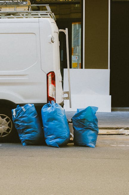 Close-up of the rear side of a white commercial van parked on a city street, with three large blue rubbish bags placed on the pavement beside it, ready for collection. The bags are made of durable plastic, slightly crinkled, and filled with household or waste materials, with some appearing more tightly packed than others. The van's rear door is partially visible, with a red tail light and a black wheel visible on the left side of the image. Behind the van, there is a building with a glass door and window, featuring a white decorative element along the bottom. The environment suggests an urban setting where private waste collection services, such as those offered by House Clearance Chelsea, might be used for rubbish removal or site clearance. The scene emphasizes the process of independent waste handling, with the bags positioned for collection or disposal, highlighting a typical setup for rubbish collection in a commercial or residential area.