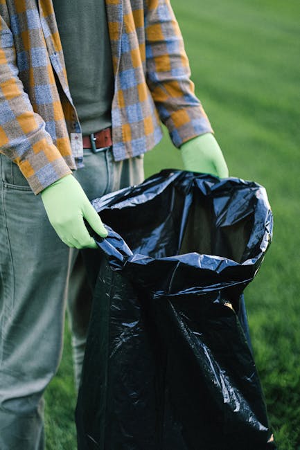 Close-up of the rear side of a white commercial van parked on a city street, with three large blue rubbish bags placed on the pavement beside it, ready for collection. The bags are made of durable plastic, slightly crinkled, and filled with household or waste materials, with some appearing more tightly packed than others. The van's rear door is partially visible, with a red tail light and a black wheel visible on the left side of the image. Behind the van, there is a building with a glass door and window, featuring a white decorative element along the bottom. The environment suggests an urban setting where private waste collection services, such as those offered by House Clearance Chelsea, might be used for rubbish removal or site clearance. The scene emphasizes the process of independent waste handling, with the bags positioned for collection or disposal, highlighting a typical setup for rubbish collection in a commercial or residential area.