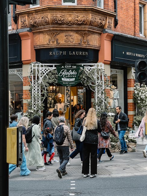 A busy street scene outside a Ralph Lauren store located within a brick building with ornate architectural details and the number 35 displayed above the entrance. The store features a decorative white metal archway with greenery and signage for Ralph's Coffee. A diverse group of pedestrians, including adults and children, are walking past and gathering near the storefront, some engaged in conversation and others passing by. The storefront window displays mannequins dressed in fashionable clothing, and floral arrangements are visible on the sides of the entrance. The surrounding environment includes a paved sidewalk with street markings, and the overall atmosphere is lively, highlighting a commercial area that may also be involved in private or independent retail activities alongside the visible clothing and accessories. The scene is well-lit, suggesting daylight conditions.