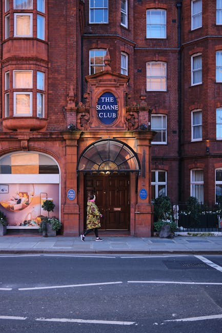 The image depicts a two-storey red brick building with white-framed sash windows and a grey tiled pitched roof, situated on a street corner. In front of the building, there is a paved sidewalk with some fallen leaves scattered across it. A black cylindrical trash bin is positioned near the edge of the sidewalk, and a white bicycle is leaning against a sign promoting private waste disposal in Chelsea. The sign, mounted on a short metal post, indicates a designated parking area and advises residents about waste collection policies. Partially visible foliage from a large leafy tree extends into the top left corner of the image, providing some greenery contrast to the urban scene. The building's exterior includes black drainage pipes running vertically along the wall, and a small window box with greenery beneath one of the upper windows. The overall scene suggests a quiet residential neighbourhood with an emphasis on independent waste management and rubbish removal services, consistent with private collection options in Chelsea.